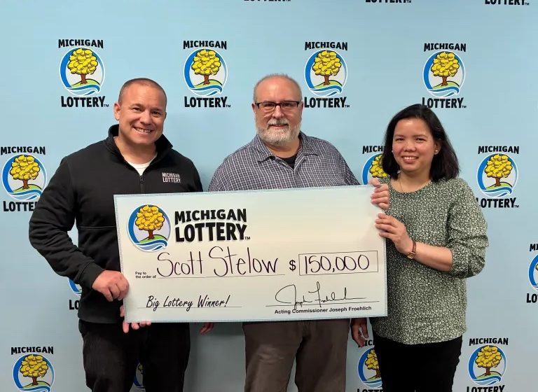 Three people hold a large Michigan Lottery check for $150,000 made out to Scott Stelow, posing in front of a backdrop covered with Michigan Lottery logos.