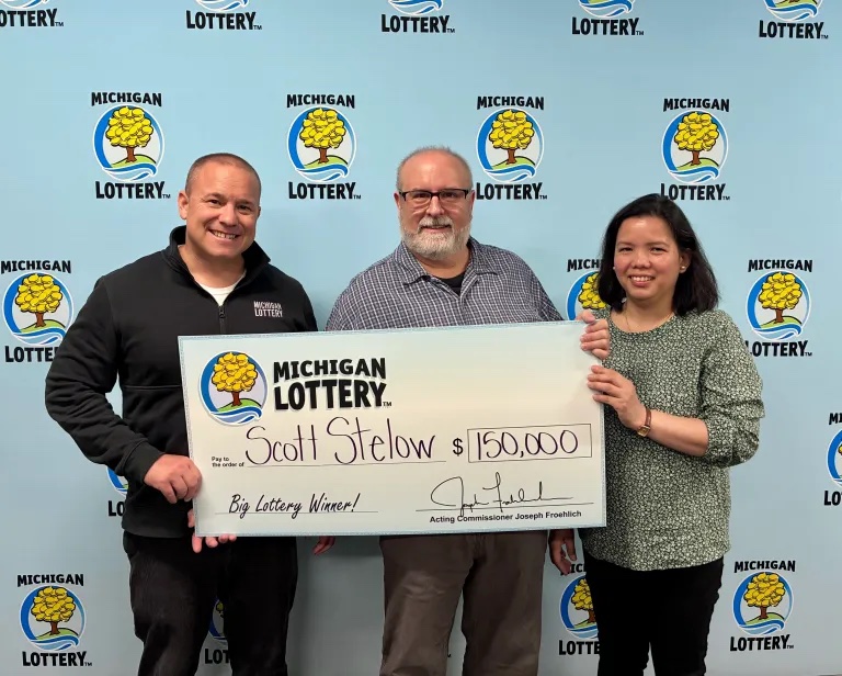 Three people hold a large Michigan Lottery check for $150,000 made out to Scott Stelow, posing in front of a backdrop covered with Michigan Lottery logos.