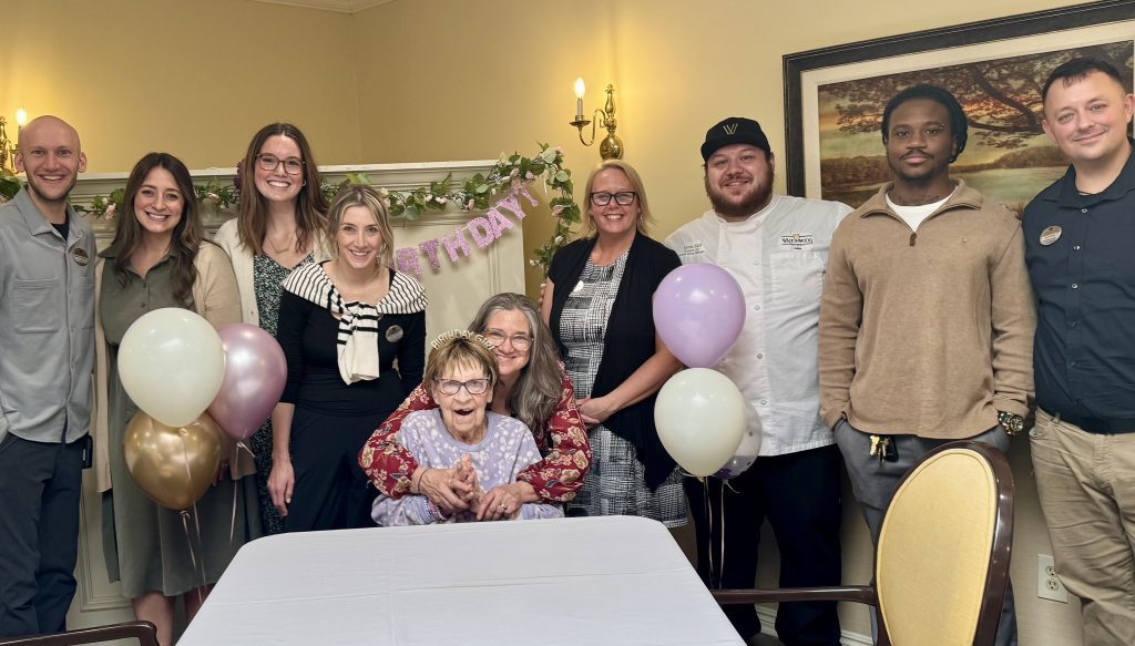 A group of adults gathered around an elderly woman celebrating a birthday, with balloons and a pastel banner in a decorated room.