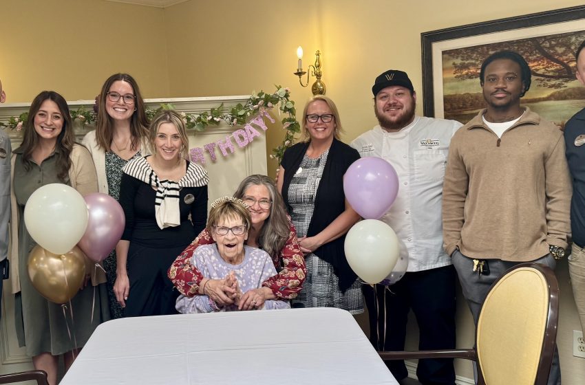 A group of adults gathered around an elderly woman celebrating a birthday, with balloons and a pastel banner in a decorated room.