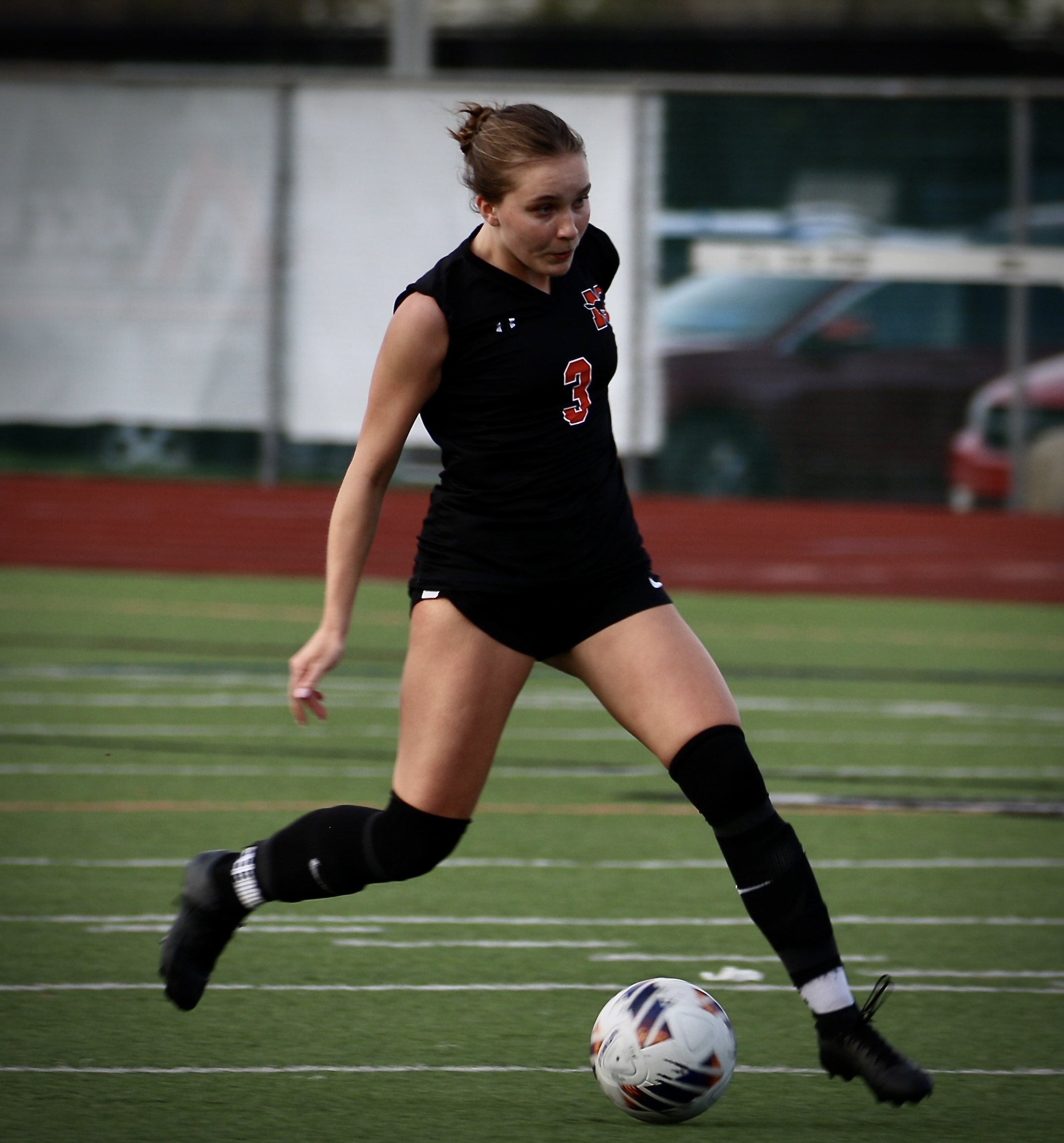 Female soccer player in a black jersey with number 3, kicking a ball on a turf field.
