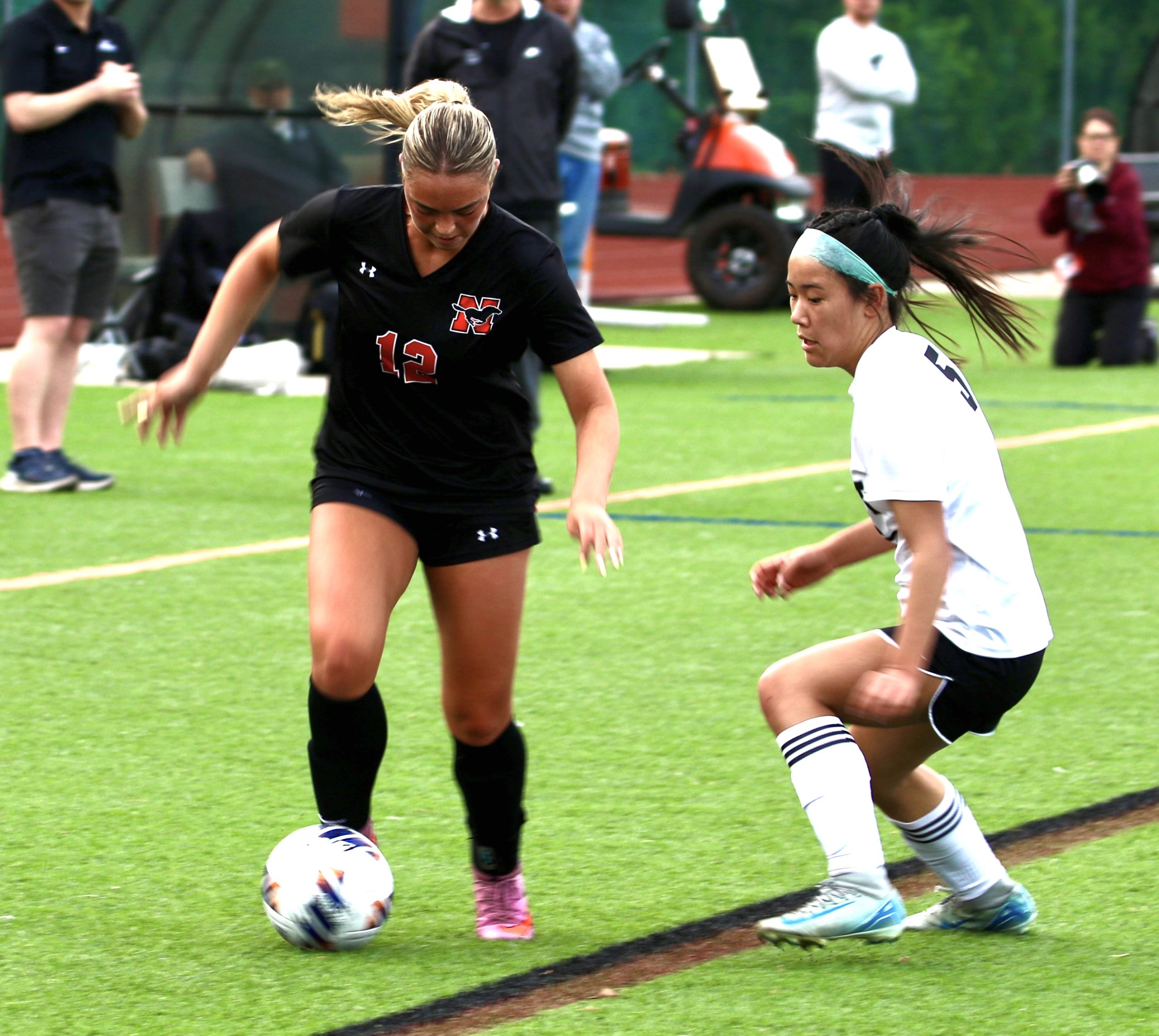 Two female soccer players compete for the ball on a green turf field, one in a black jersey (no. 12) controlling the ball while the other in white defends closely.