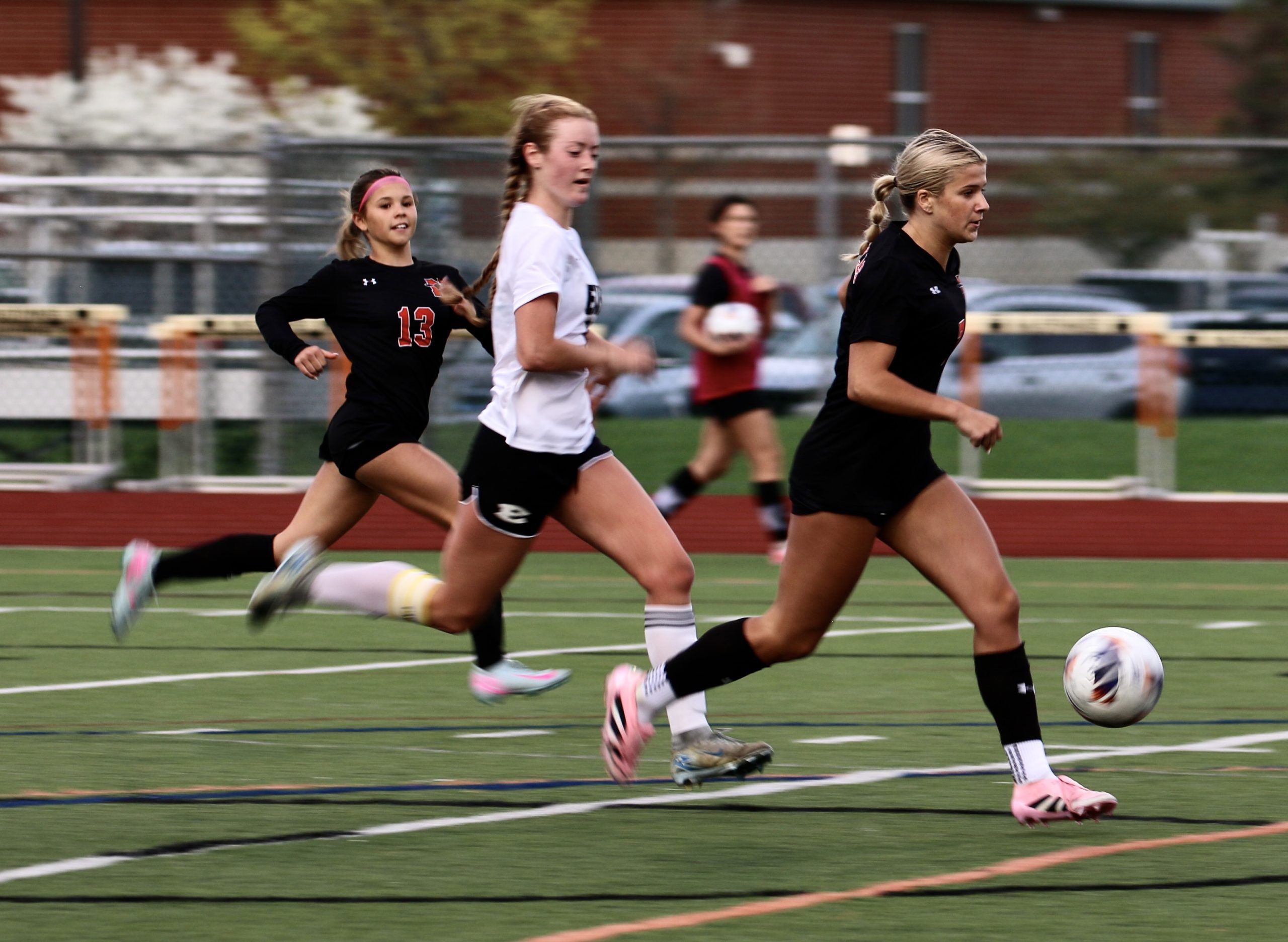 Female soccer players sprint on a green field as a ball rolls ahead, with one player in black leading.