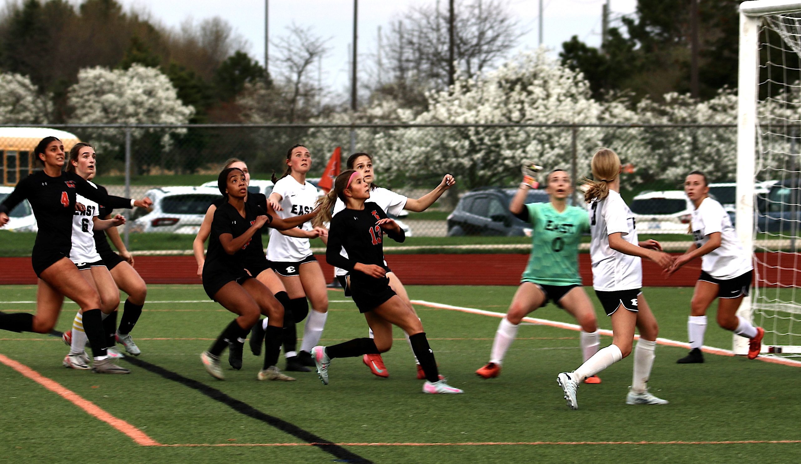 Girls' soccer match with players in black and white uniforms racing toward the goal on a turf field, goalkeeper in green nearby.