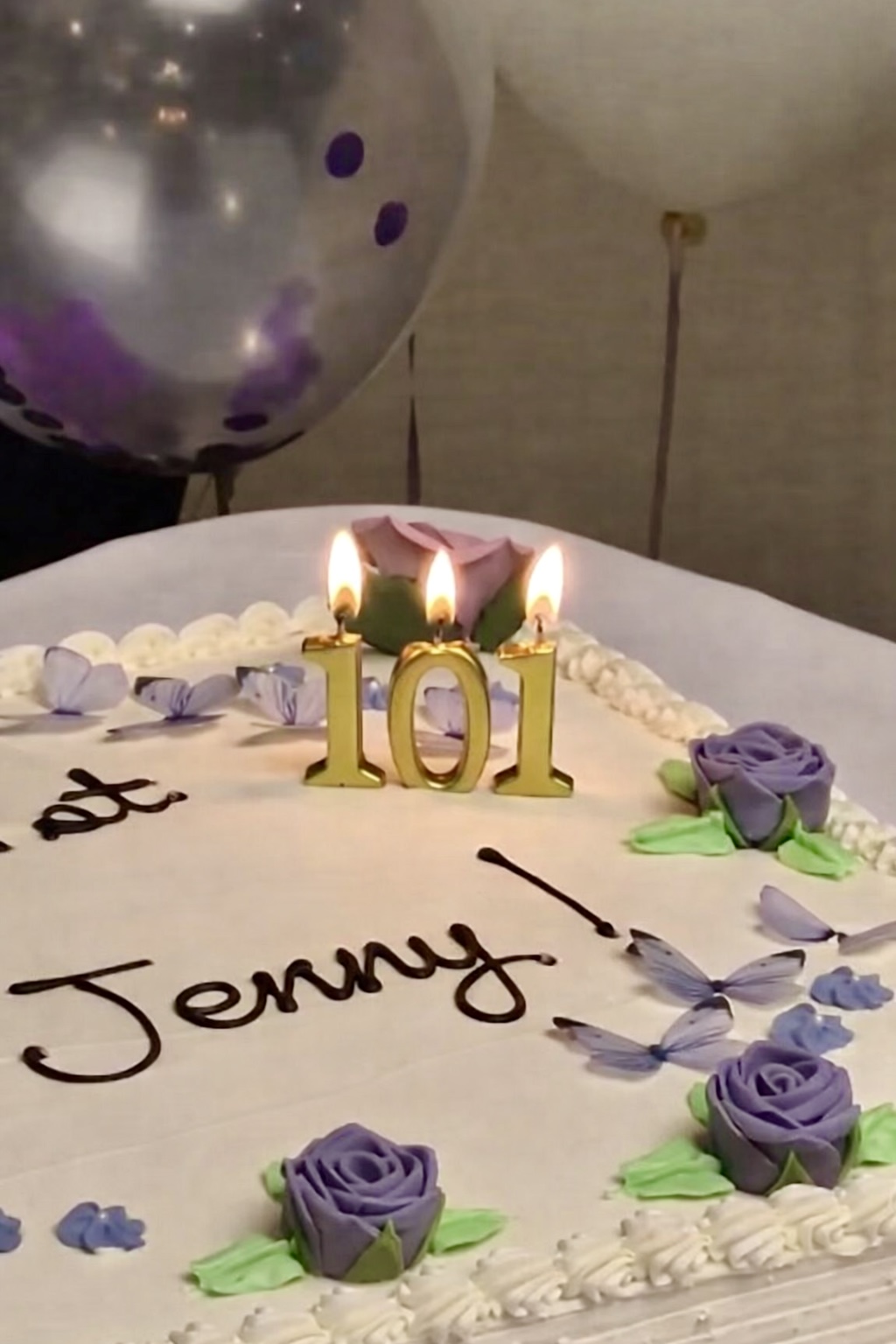 Birthday cake with lit gold '101' candles, purple silk roses, and the name Jenny written in icing beside blue butterfly decorations.