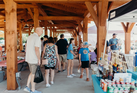 People browse vendor stalls under a wooden outdoor market pavilion; a man carries a black bag while a group gathers near a beverage display and a boy in a blue shirt nearby.