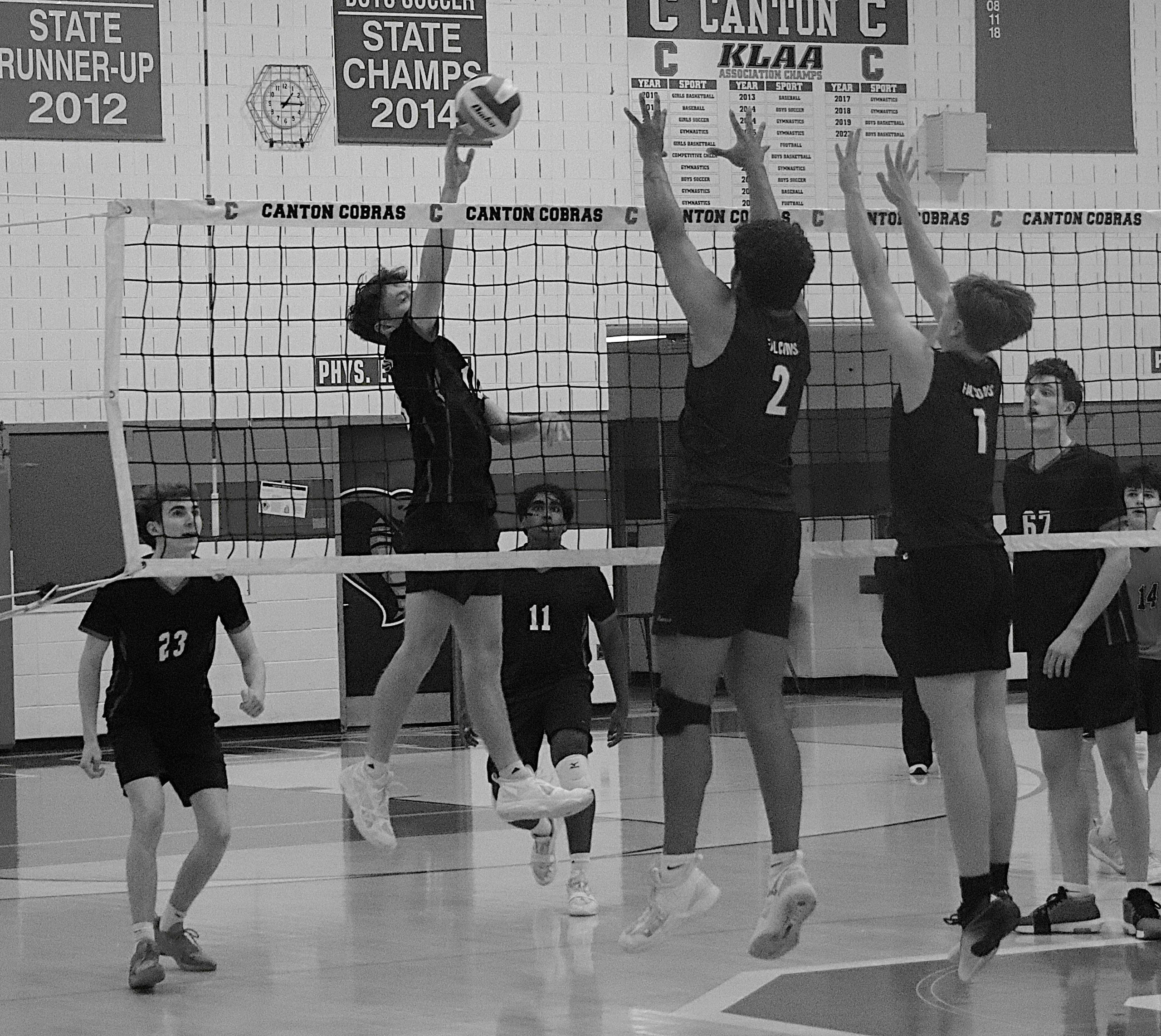 Girls' volleyball game in a gym; players leap at the net as the ball heads over, banners on the wall read state champions 2012 and 2014.