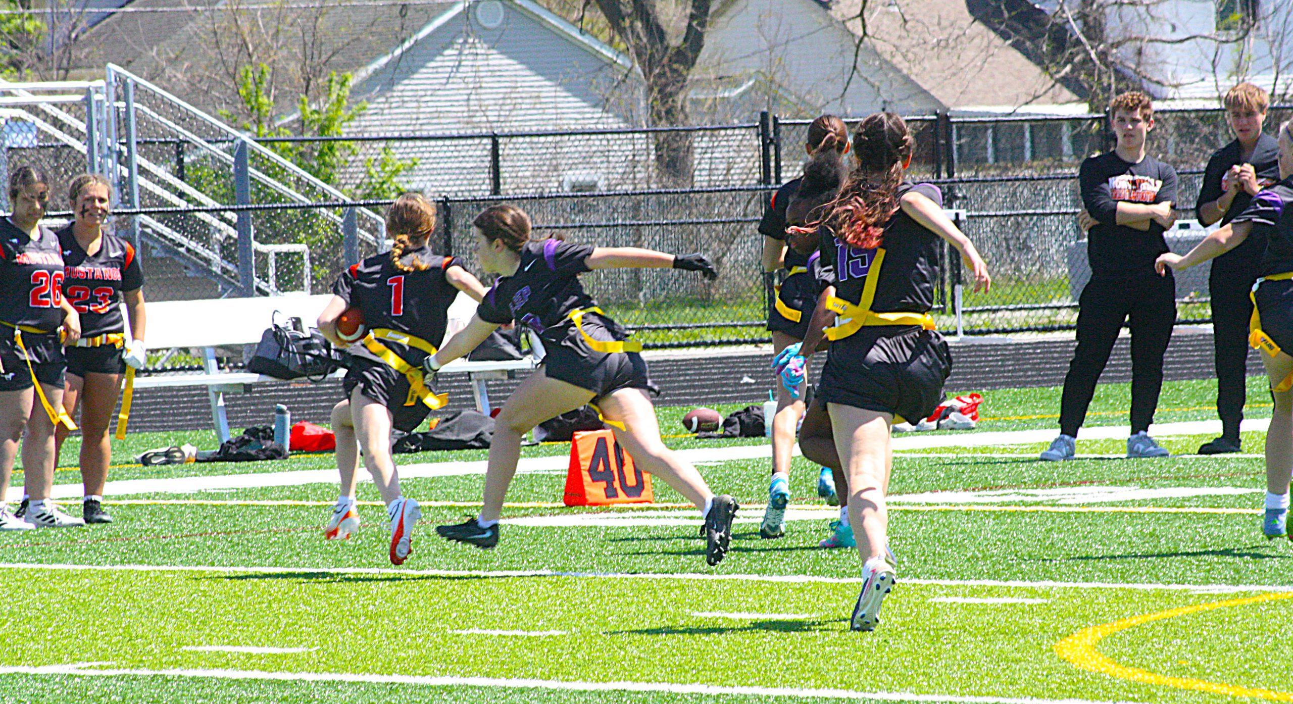 Girls’ flag football game on a bright artificial turf field; several players in black uniforms with yellow flags race toward the ball while a few teammates and spectators watch near a chain-link fence.