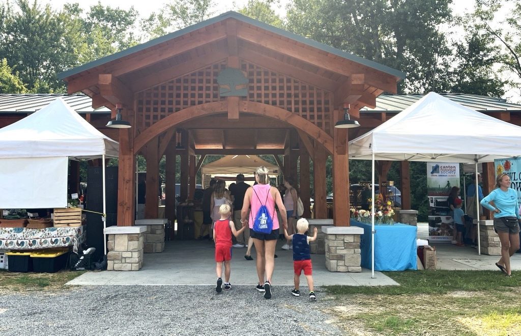 Family walks toward a wooden pavilion with white canopy tents at an outdoor market. A woman in a pink backpack holds two young children's hands as they approach the structure.