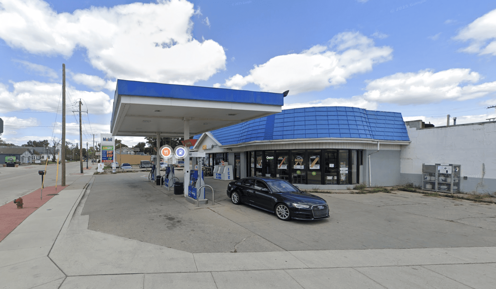 Gas station with a blue curved canopy and convenience store; a black Audi parked near the pumps.