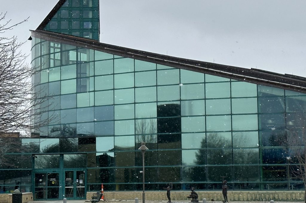 Glass-fronted modern building with a tall triangular peak, facing the camera, in a light winter scene with snow on the ground and pedestrians nearby