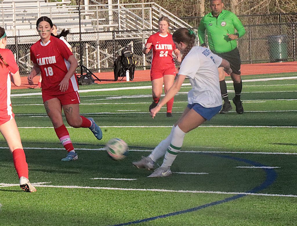 Youth soccer game with players in red Canton uniforms chasing the ball on artificial turf, a player in white nearby, and a green-clad referee in the background.