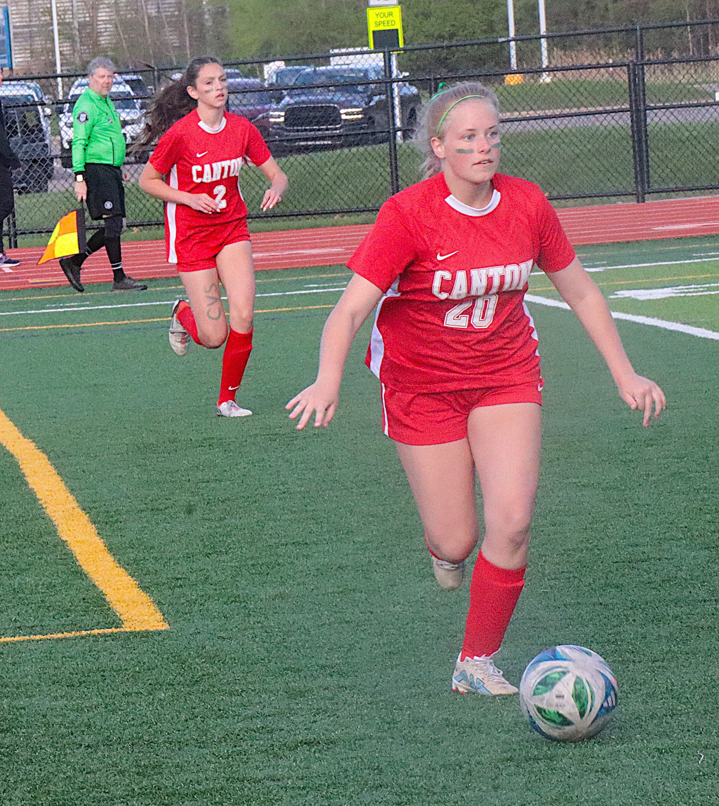Two female soccer players in red uniforms sprint on a turf field with a ball near the foreground; player in front wears number 40, another behind wears number 2