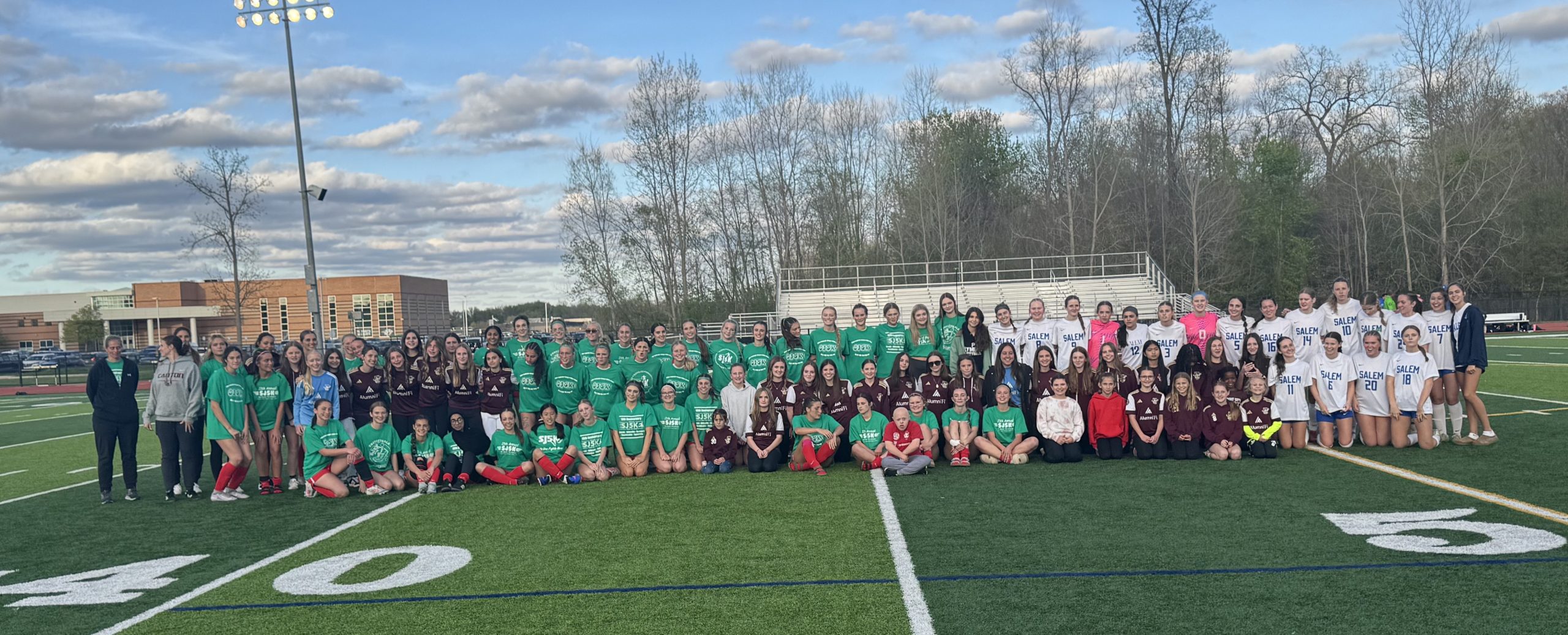 Large group of students posing for a team photo on a football field, wearing green, white, and maroon jerseys against a backdrop of bleachers and trees.