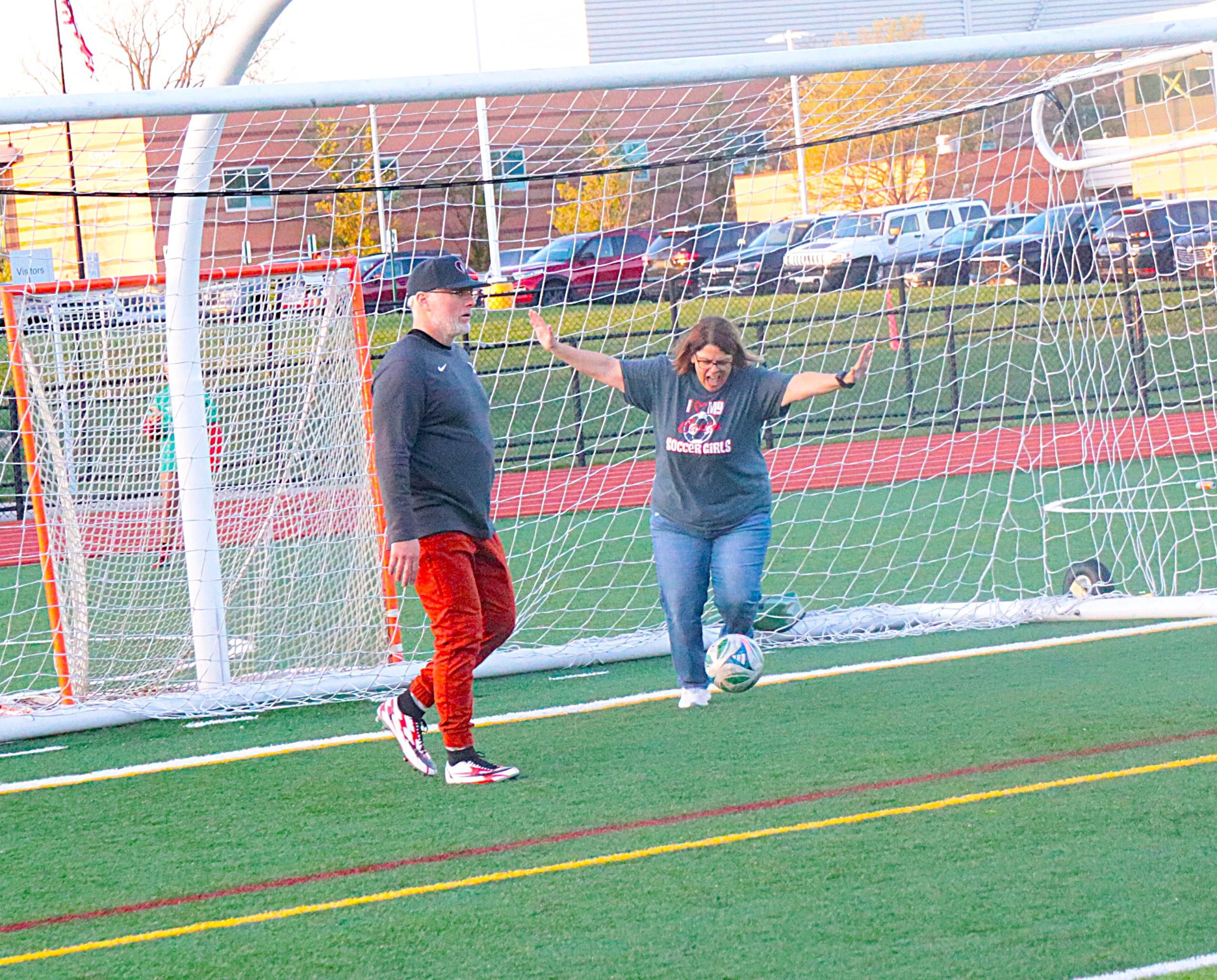 Two adults by a soccer goal on an artificial turf field; a man in black and red pants stands beside a woman in a gray shirt with arms raised, cars in the background.