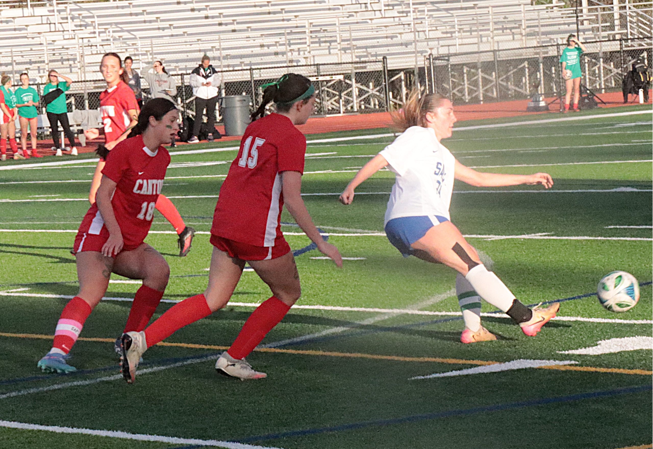 Girls' soccer game in progress: red team near the ball while a white-uniformed player kicks it on a green artificial turf field with empty bleachers in the background.