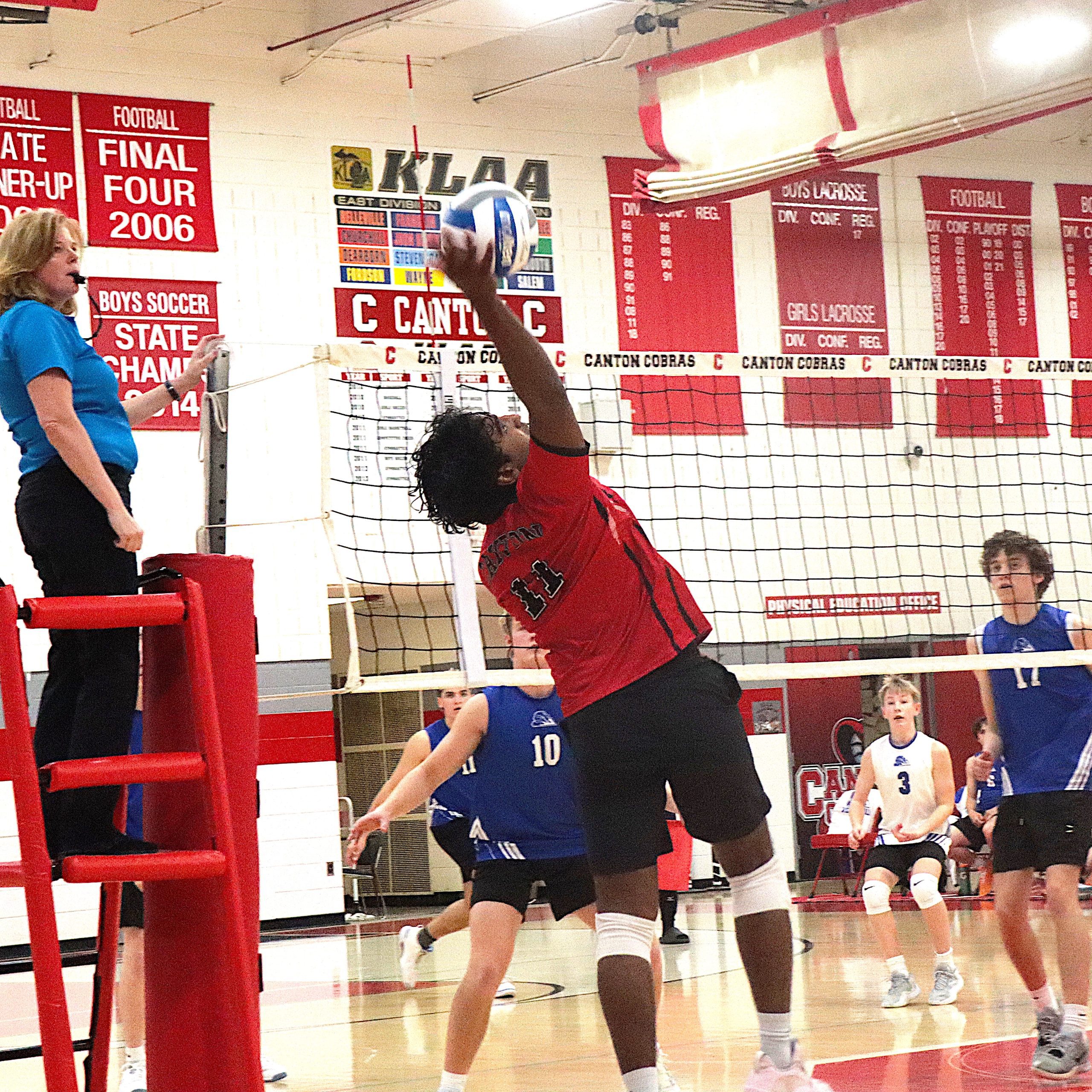 Female referee or coach in blue shirt watches as a player in red jumps to spike the ball at the net in a gym with red bleachers and banners behind.
