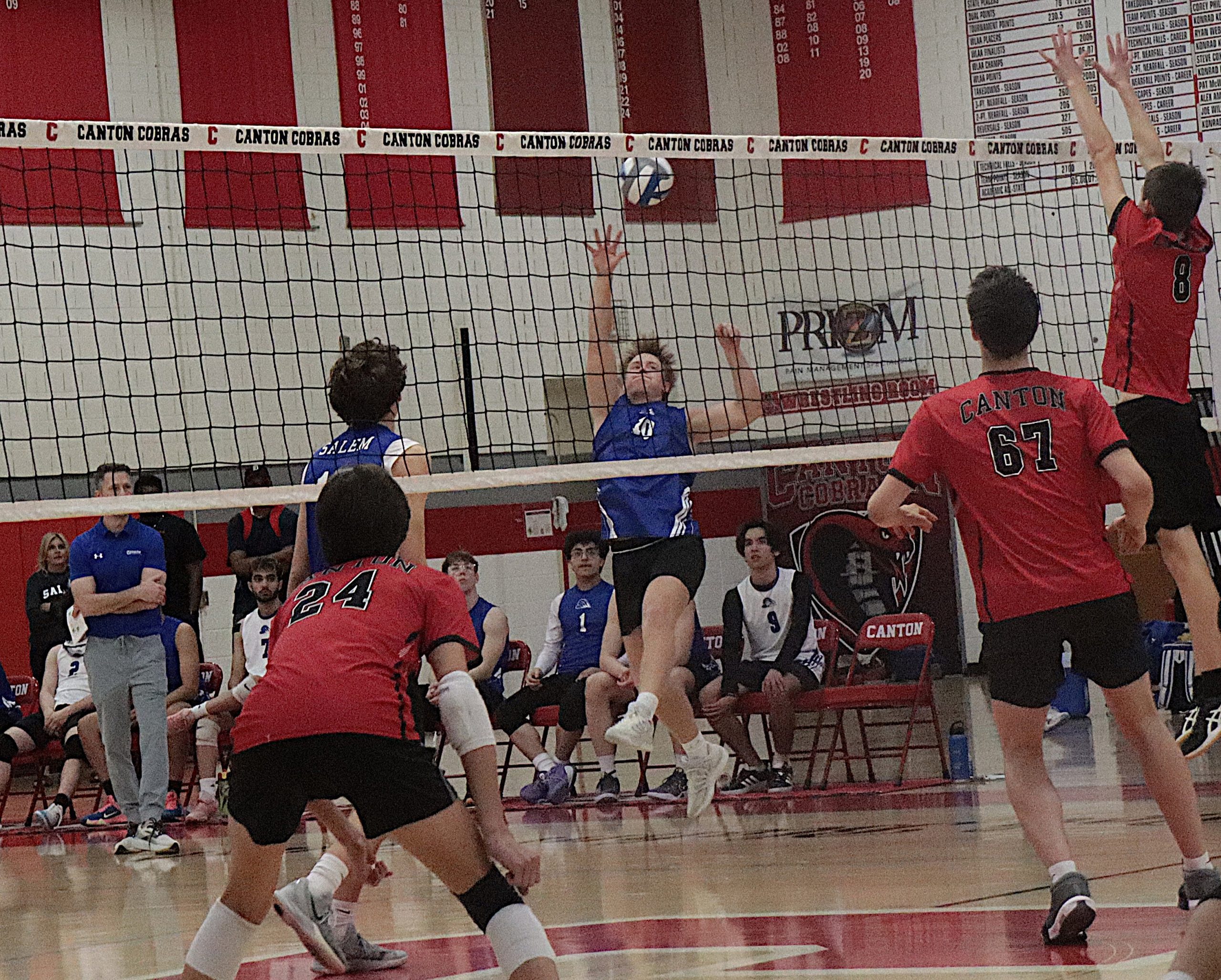 Volleyball players in red and blue uniforms jump near the net as a ball sails over, with a scoreboard in the gym background.