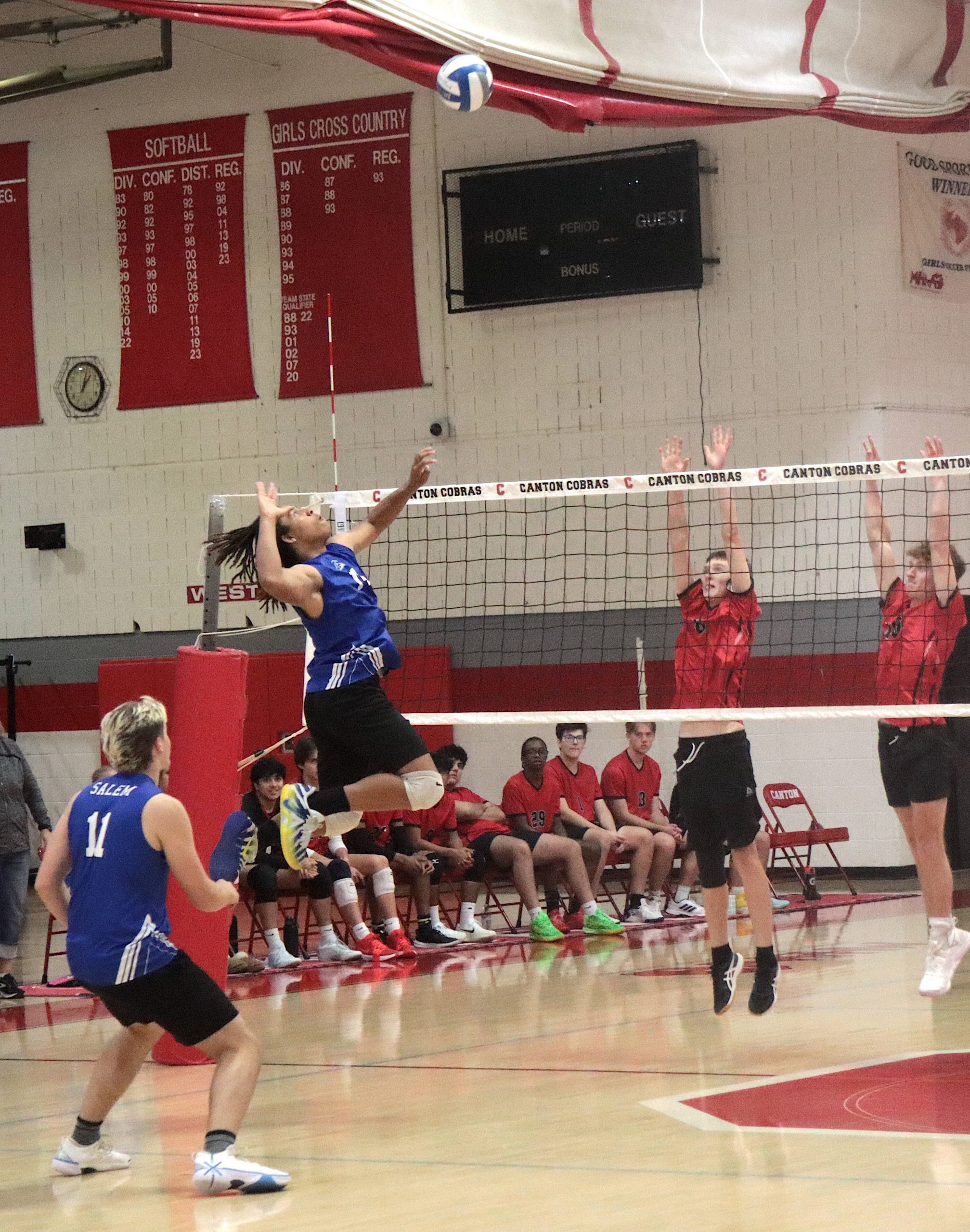 Blue-clad volleyball player mid-air near the net preparing a spike while red-team blockers reach up; bench players watch in background.