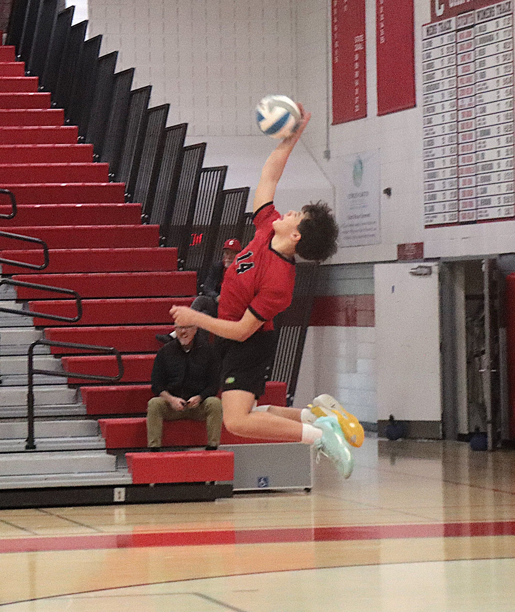 Volleyball player in a red jersey reaches up to hit a blue-and-white ball near red bleachers in a gym; spectators sit on the steps in the background.