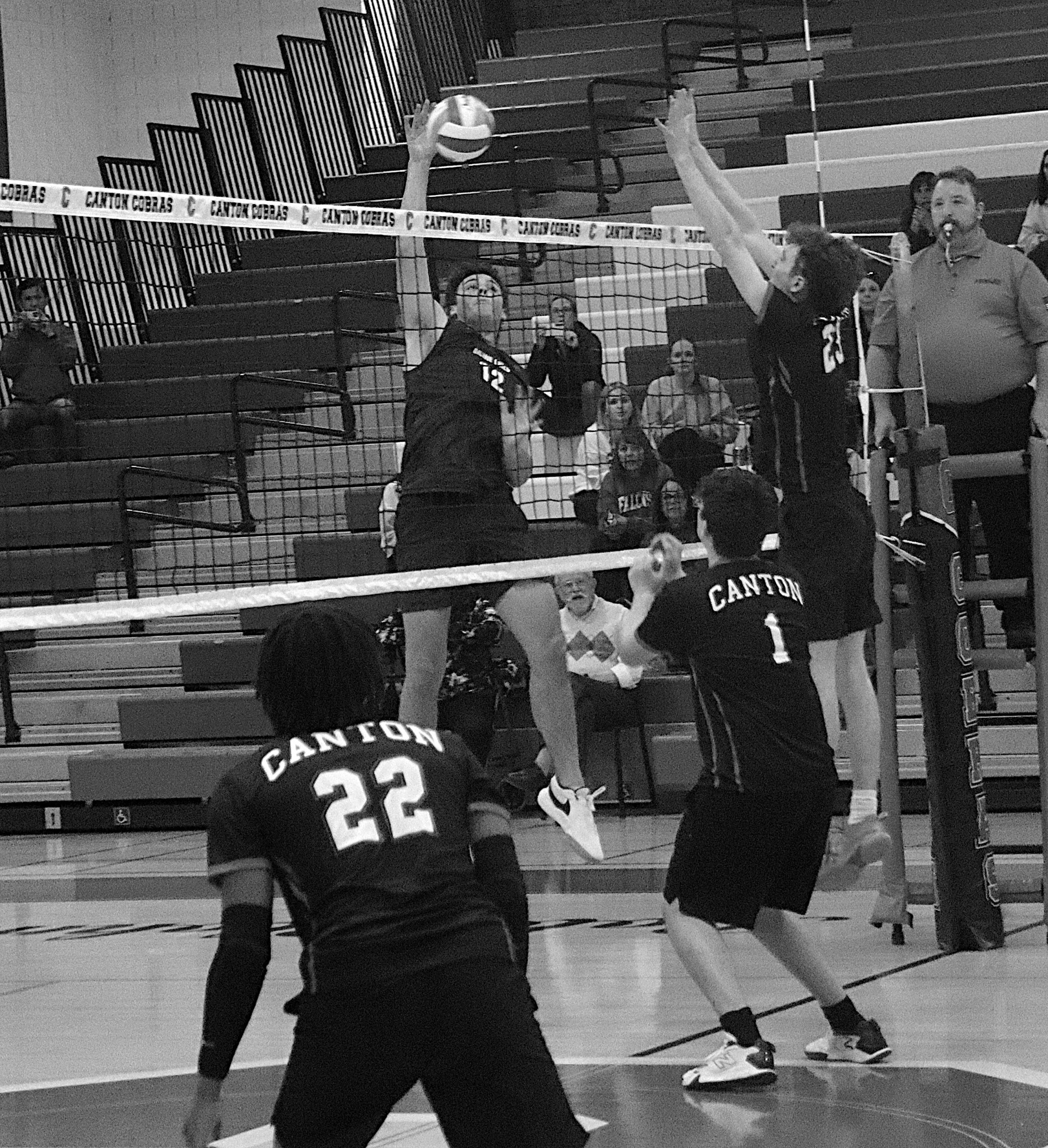 Two volleyball players jump at the net to set or spike the ball during a match in a gym, Canton jerseys visible and spectators watching.