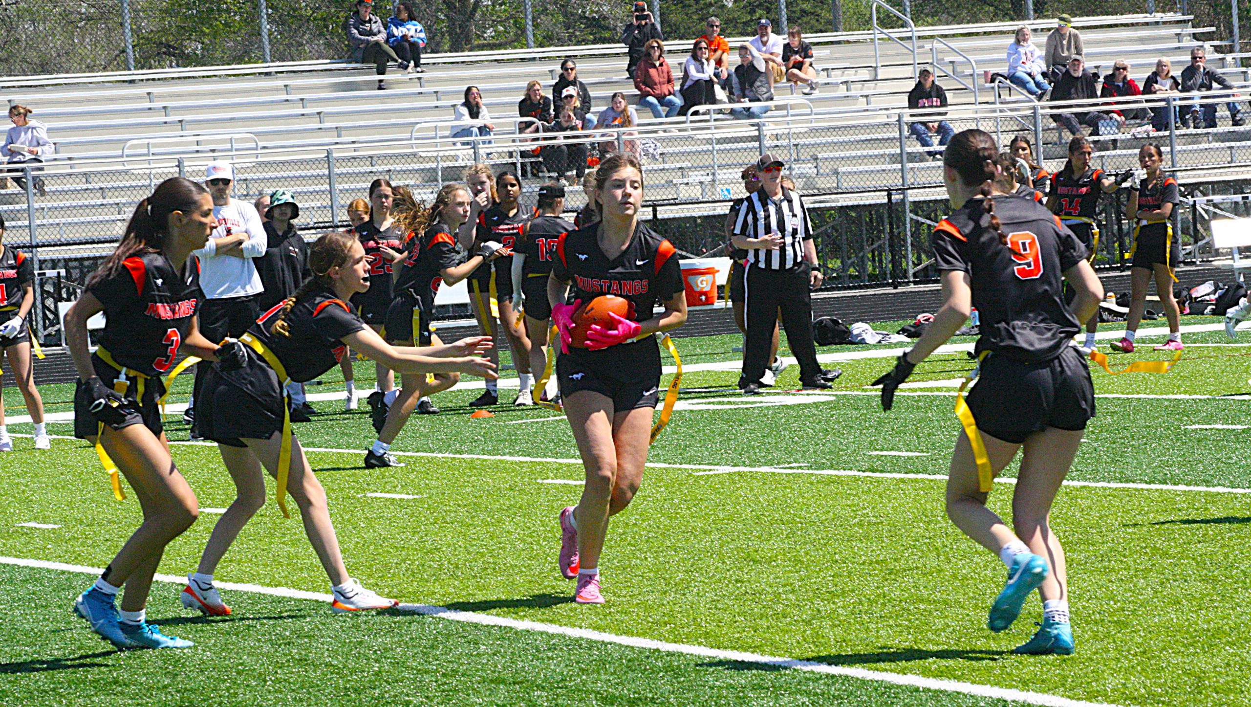 Girls' lacrosse game on a sunny day with players in black uniforms and yellow flag belts sprinting on a green turf field, spectators in bleachers in the background