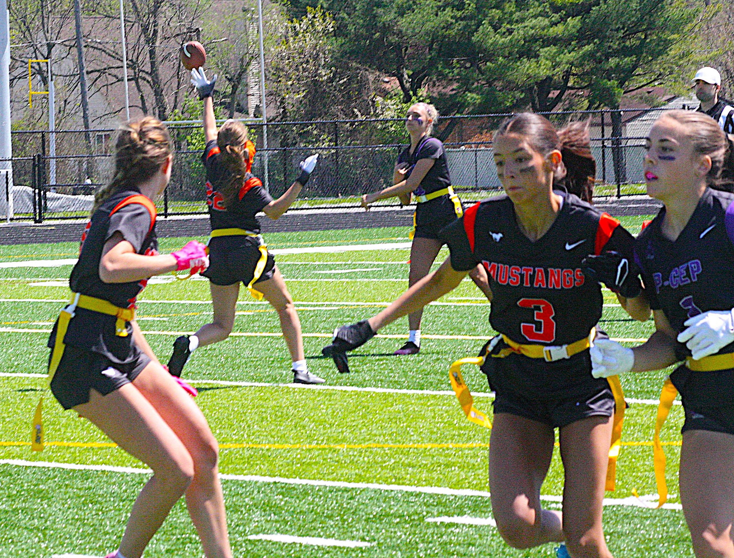 Girls in black and orange flag football uniforms sprint on a sunny field, one reaching for a ball in midair as teammates close in.