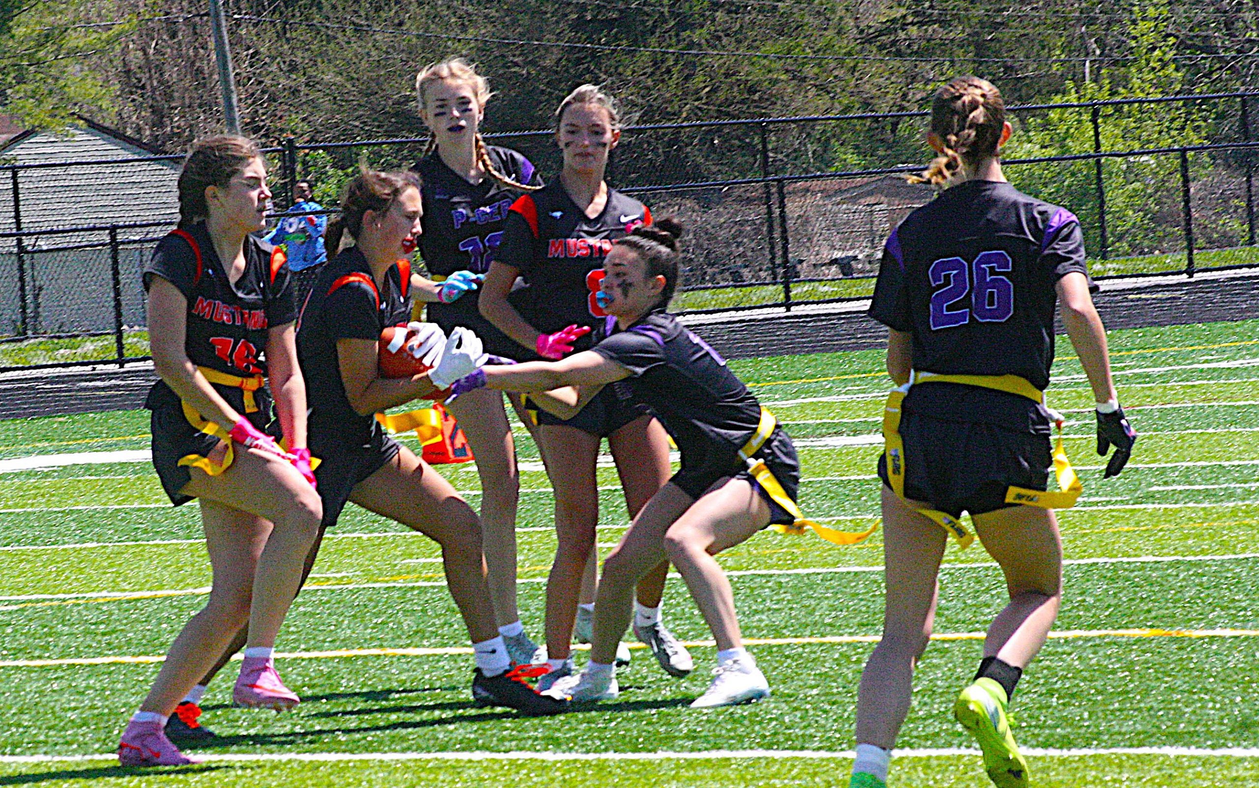 Girls' flag football team on a sunny field, wearing black jerseys with red trim and yellow flag belts, mid-game action.