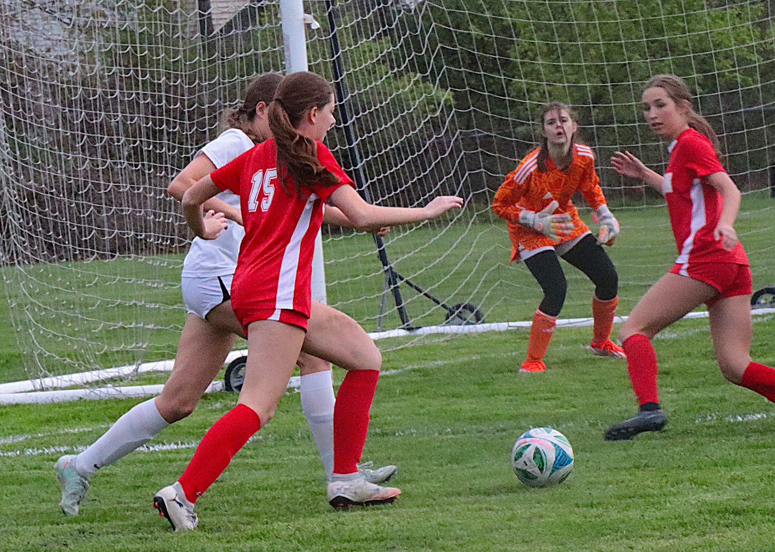 Girls' soccer match near the goal line as players chase the ball on the grass field behind the net. (informative)