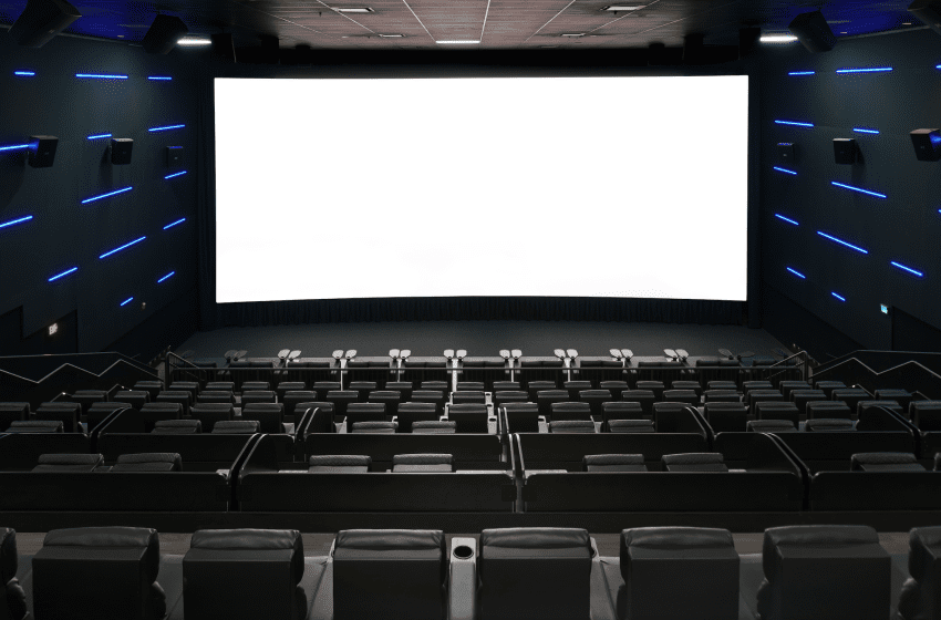 Empty modern cinema with a large white screen, dark walls, and blue LED strips along the sides above the seats.