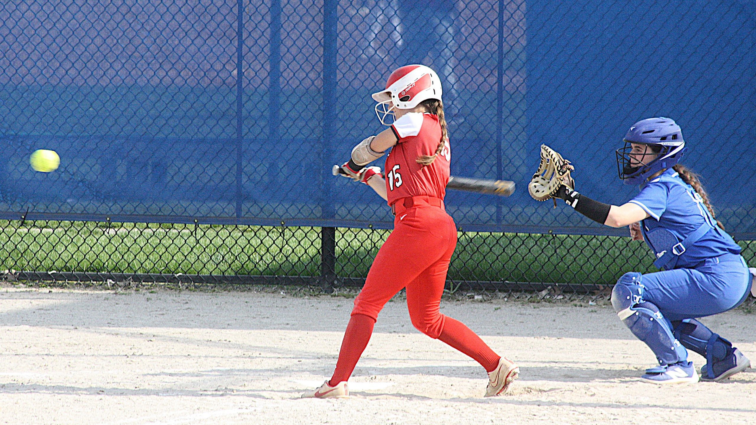 Two female softball players in a fenced field: a red-uniform batter swings at a yellow ball as a blue-uniform catcher crouches behind.