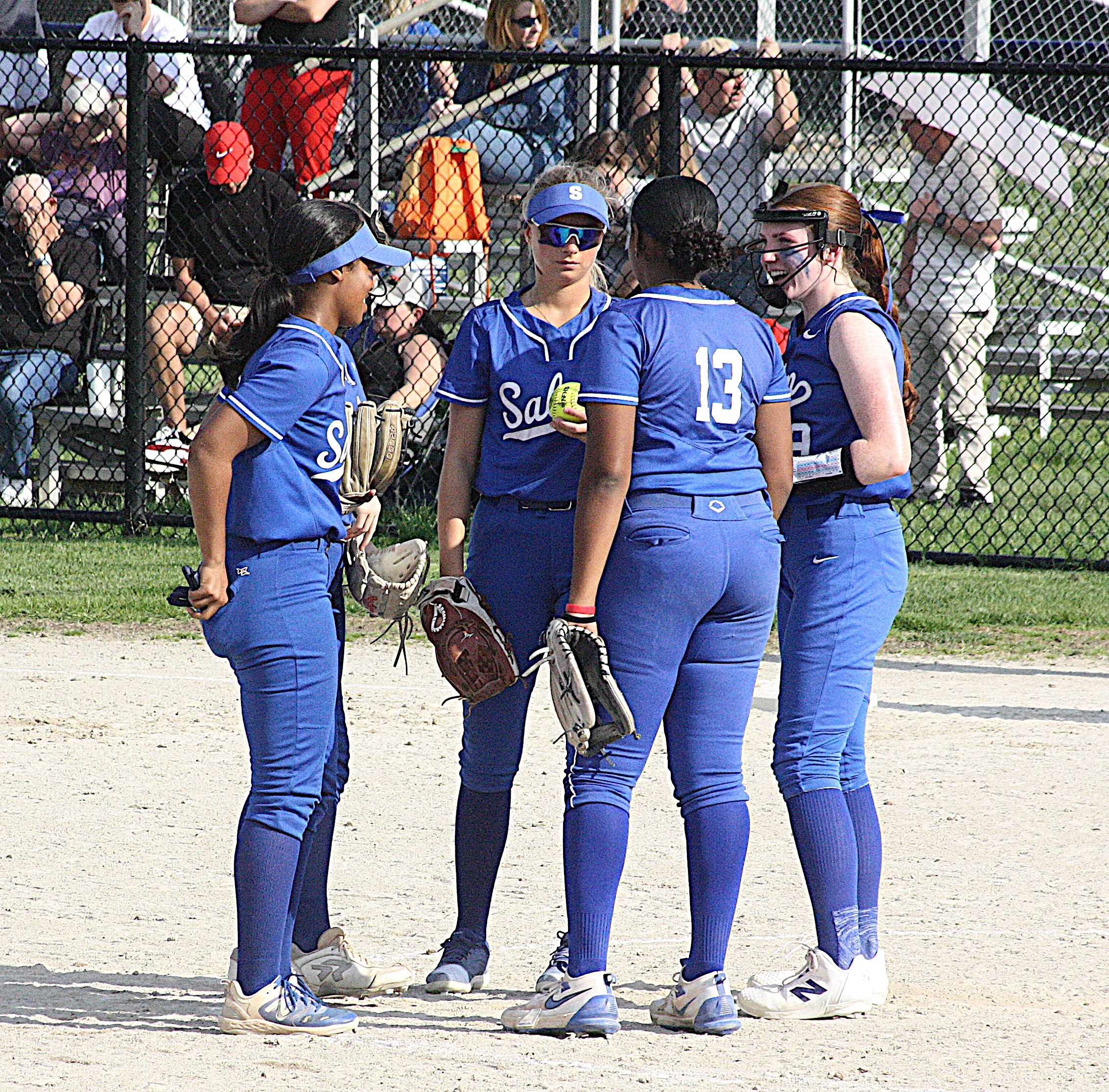 Softball players in blue uniforms stand together on a dirt field near a chain-link fence, wearing gloves and helmets.