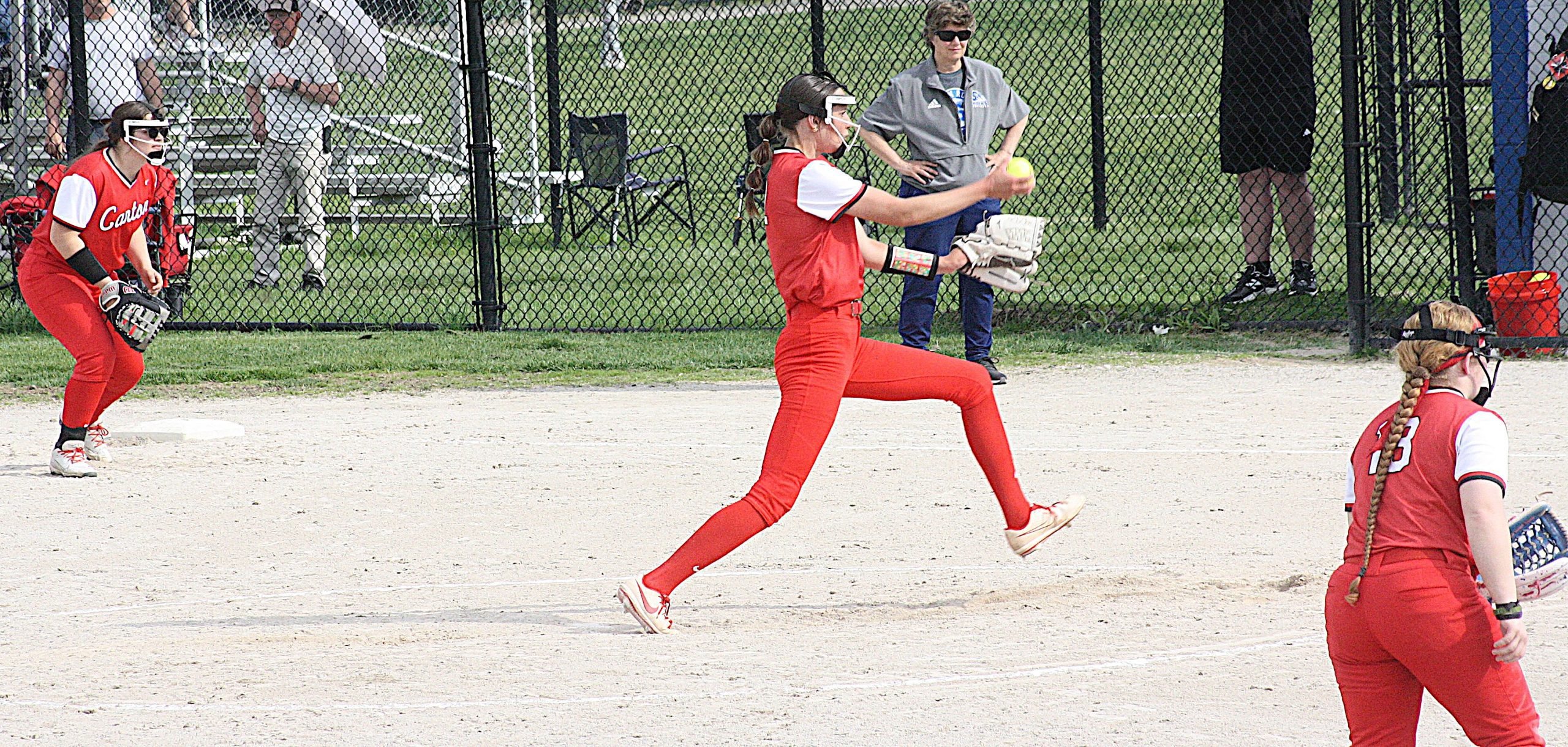 Softball pitcher in a red uniform mid-pitch on a dirt field, catcher ready behind home plate with a glove and mask in view.