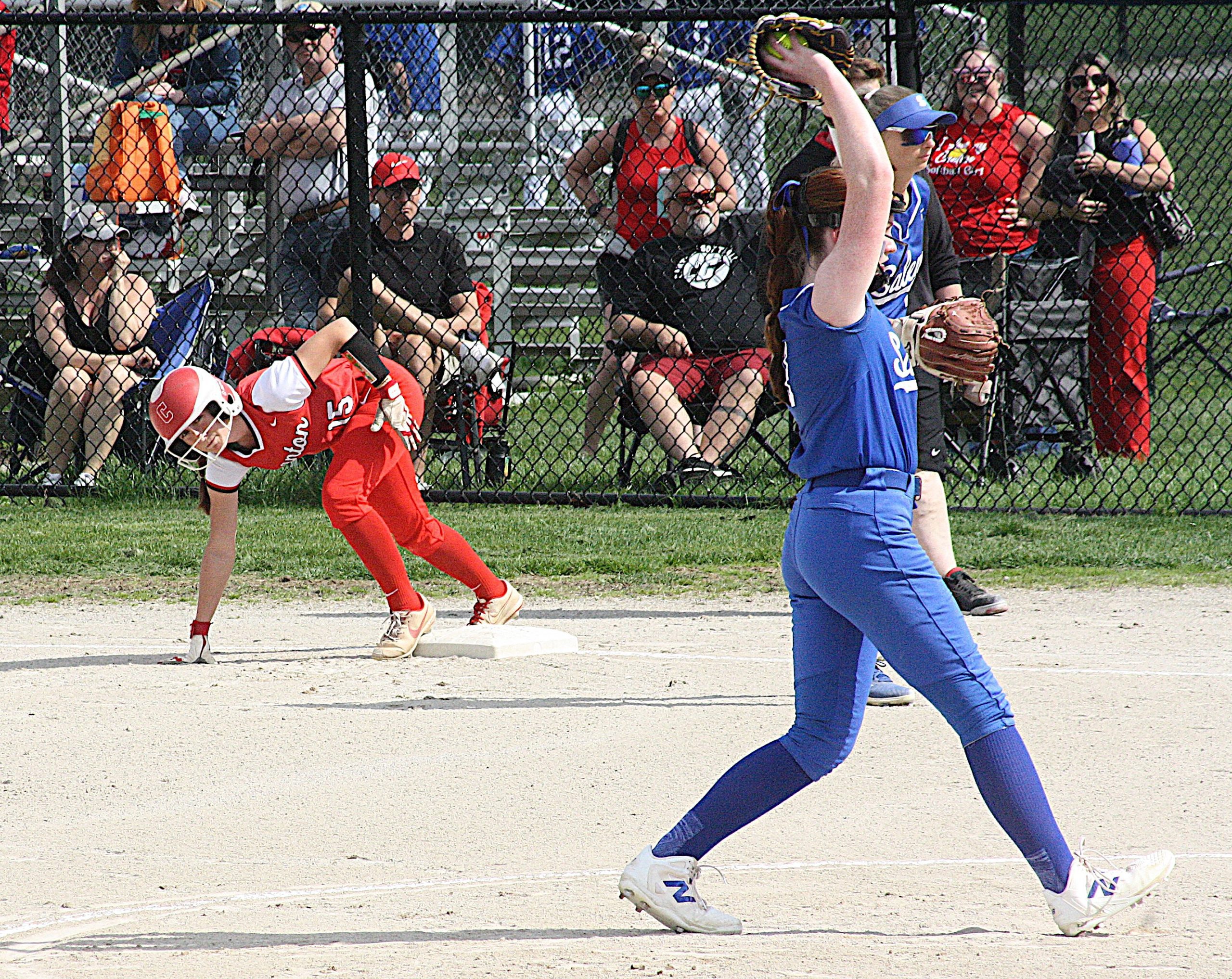 Soccer? No—softball/baseball action: a player in red slides into a base as a blue-uniform fielder reaches with a glove, near a chain-link fence with spectators behind it.