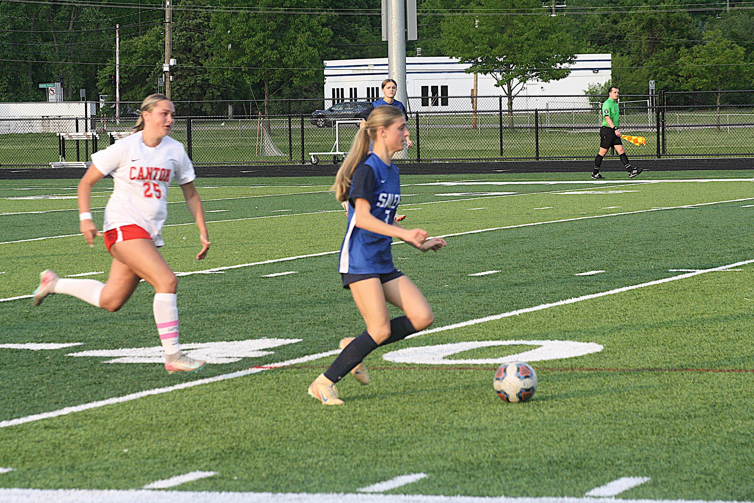 Alik3 - Social House News Soccer scene with players running on a turf field; a white jersey player (#25) ahead left, a blue jersey player in foreground, and a green-clad referee in the background.