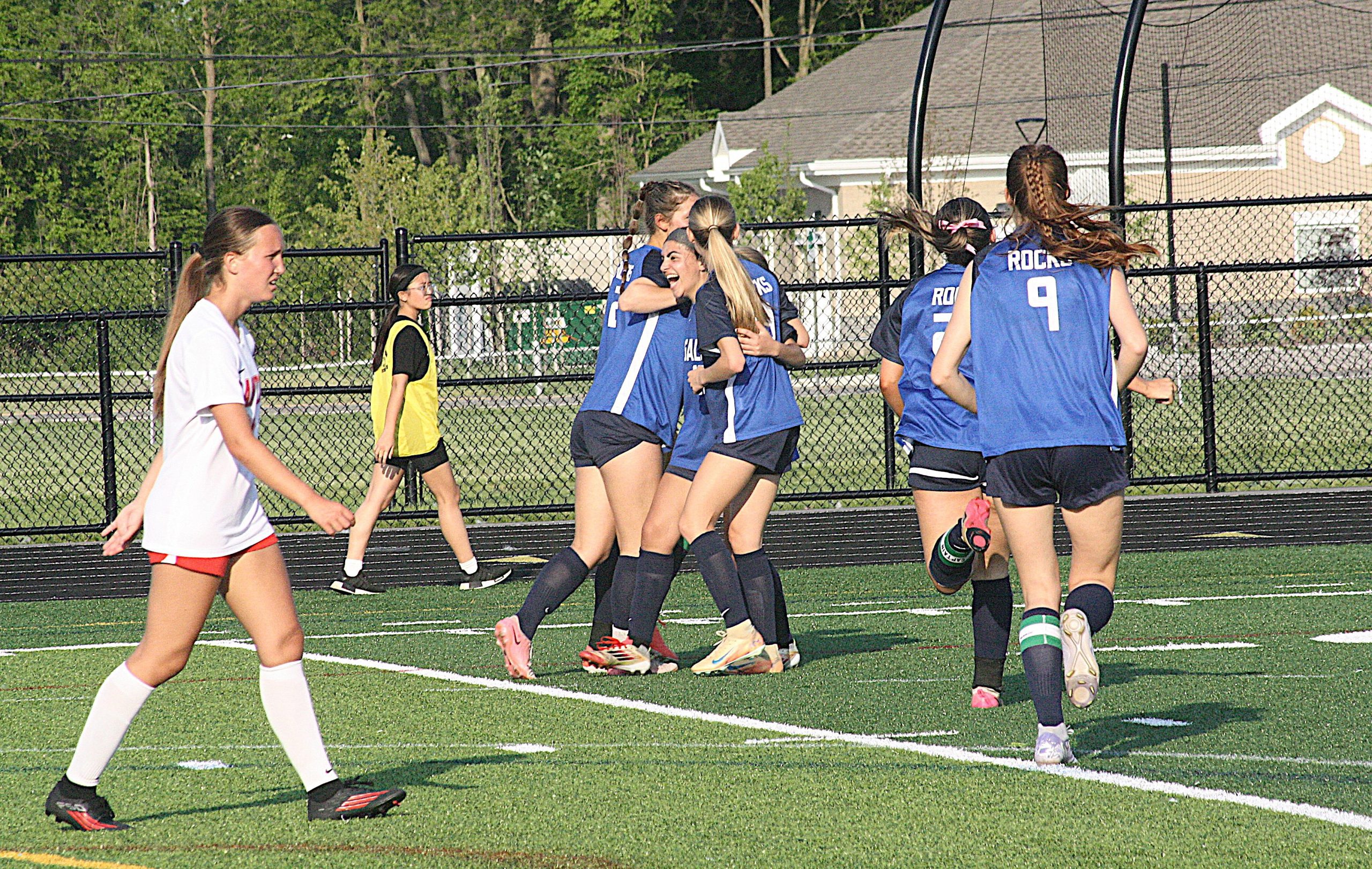 Alik4 - Social House News Girls' soccer teammates in blue jerseys celebrate and hug on an outdoor turf field near a chain-link fence.