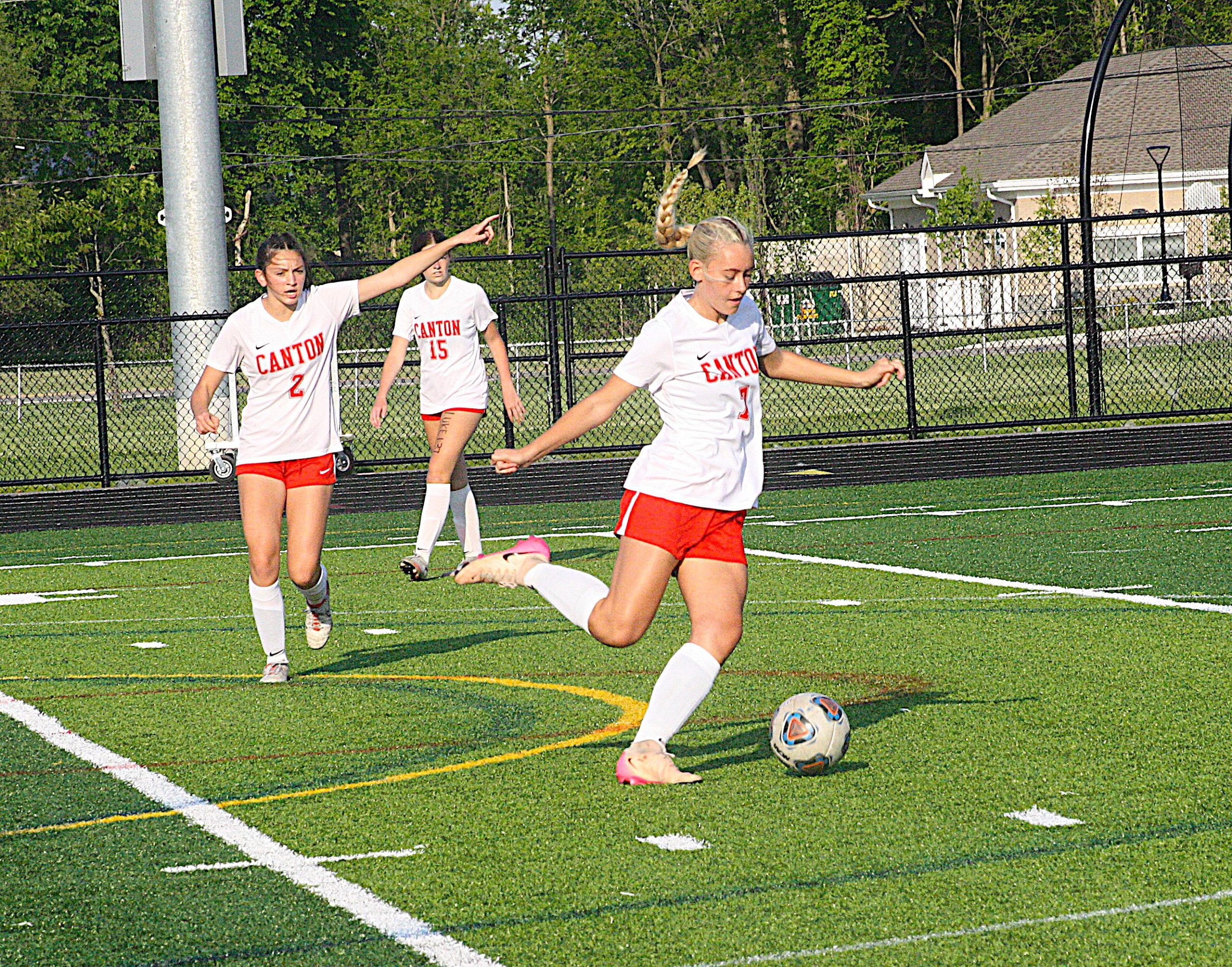 Alik6 - Social House News Girls soccer game on a fenced turf field; players wear white Canton jerseys and red shorts, ball at the foreground.