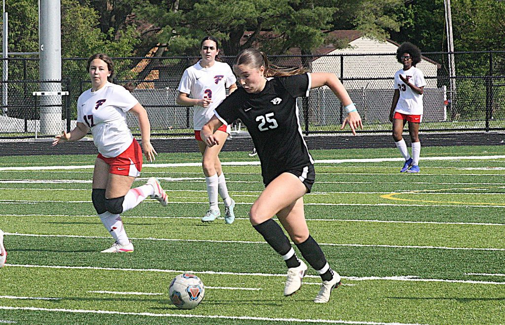 Girl in black uniform dribbles soccer ball while pursuing play, with teammates in white jerseys nearby on a sunny field.