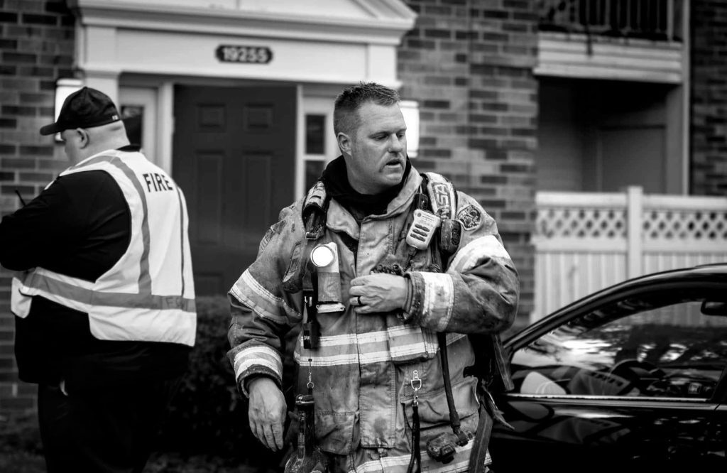Firefighter in full turnout gear stands outside a brick home, talking to a colleague wearing a reflective vest, at an emergency scene.