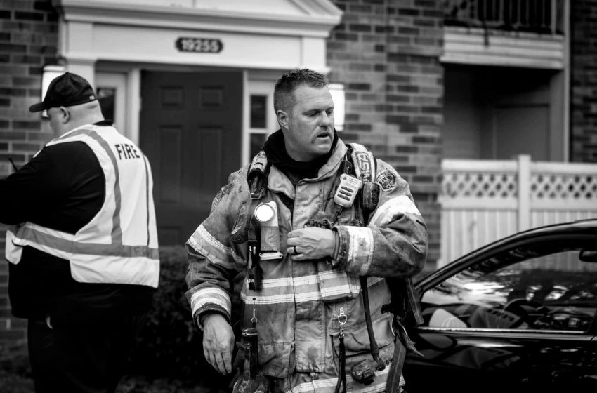 Firefighter in full turnout gear stands outside a brick home, talking to a colleague wearing a reflective vest, at an emergency scene.