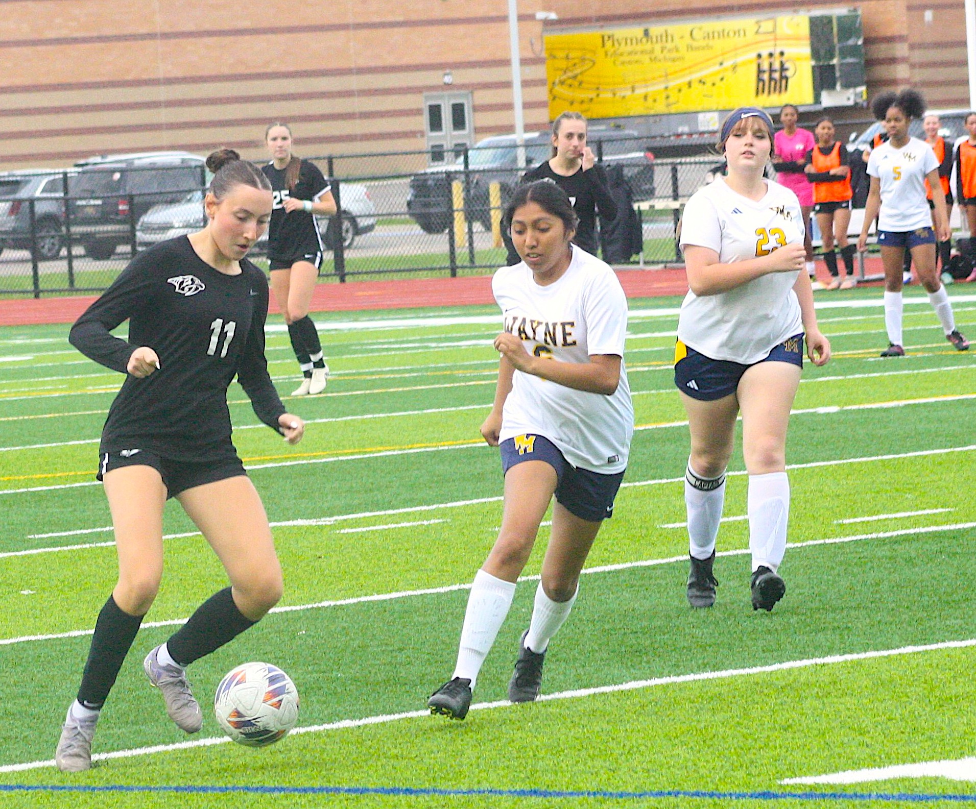 Girls' soccer players chase the ball on a bright green field during a match; a player in black leads ahead of others in white uniforms with yellow numbers on the right.