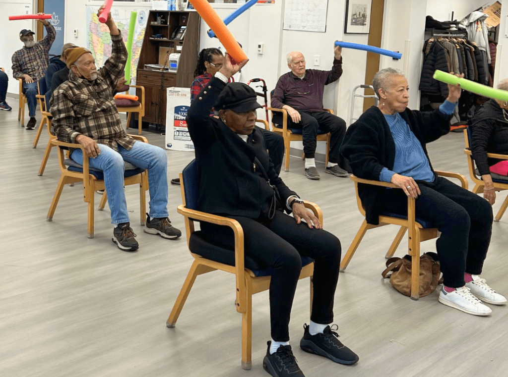 Group of seniors seated in a circle performing seated arm stretches with colorful foam sticks som connected environment in a community center gymroom.