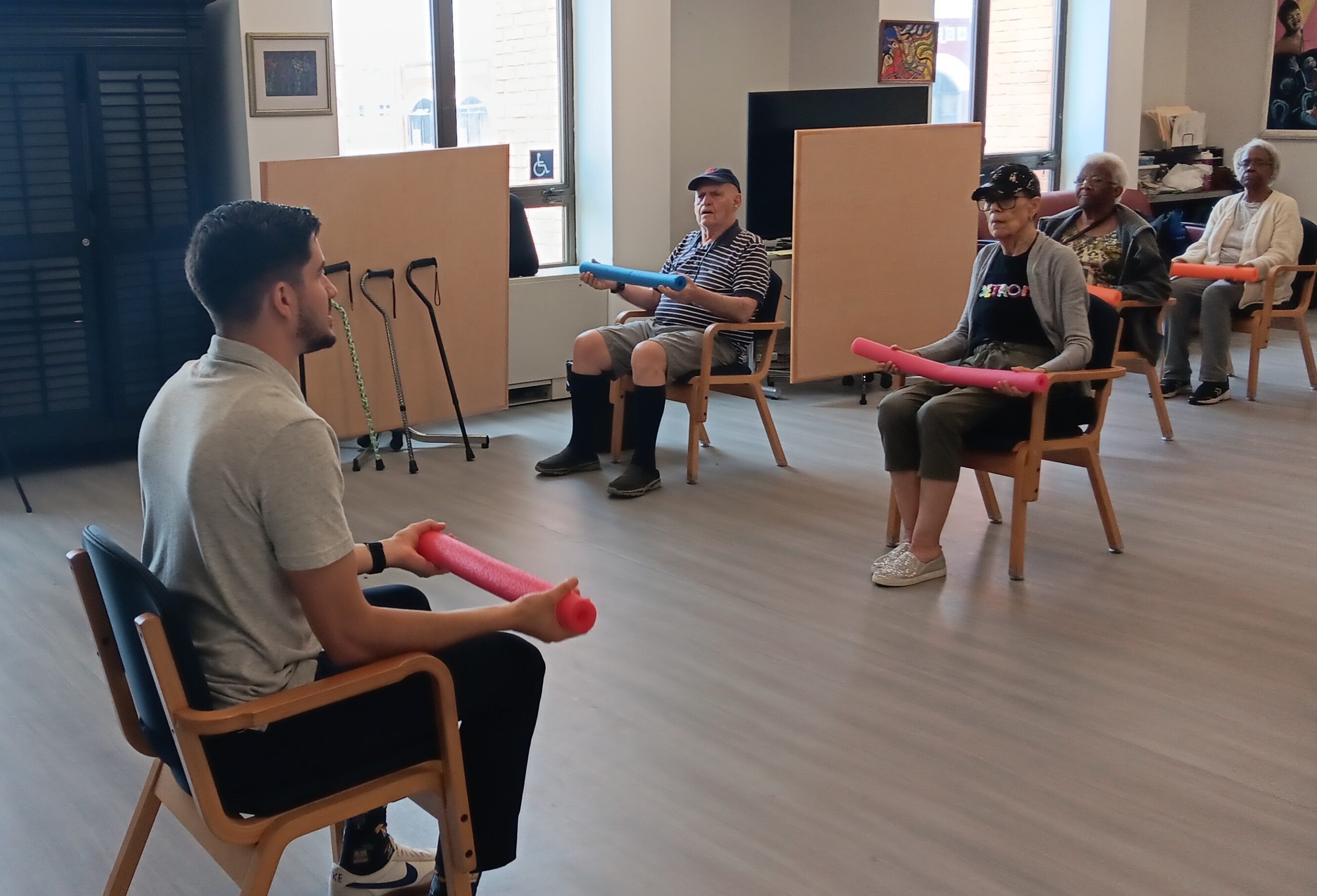 Seniors seated in chairs holding colorful foam exercise noodles during a chair workout in a bright room.