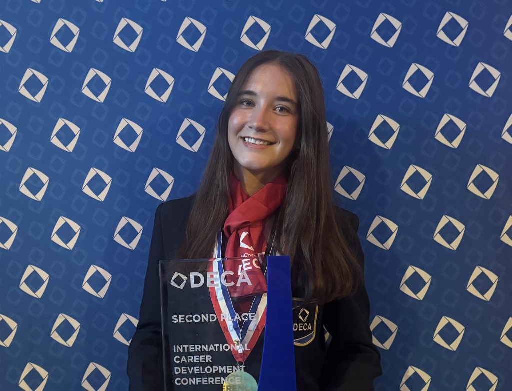 Smiling young woman in a blazer and red scarf poses with a glass award in front of a blue DECA backdrop. The plaque reads 'Second Place' at the International Career Development Conference.