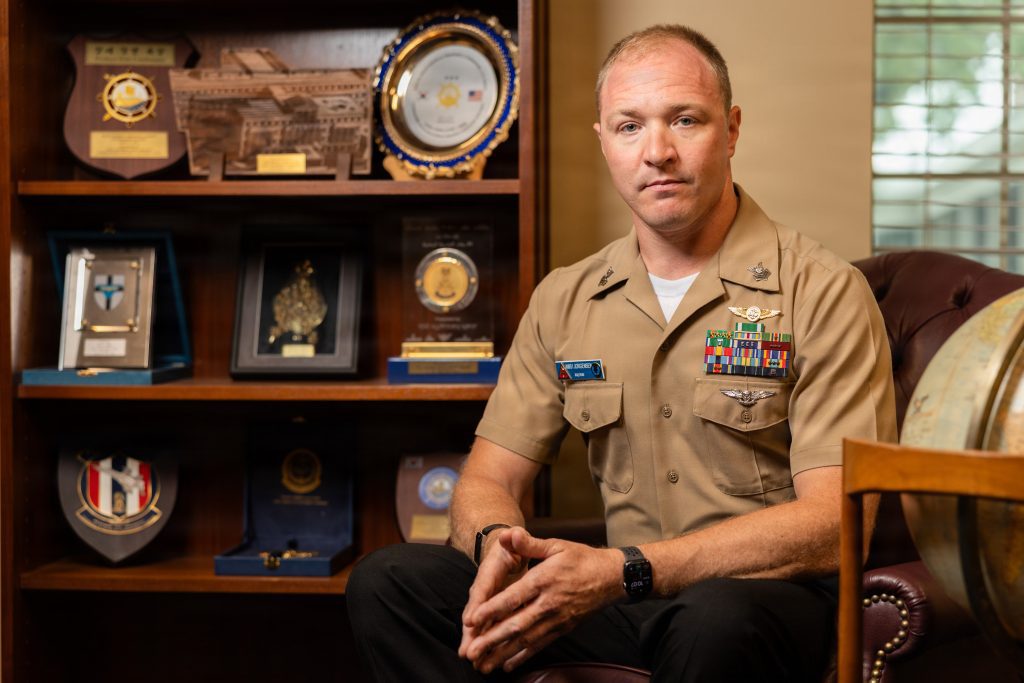 Military officer in a tan dress uniform sits in front of a shelf of awards and plaques, facing the camera.