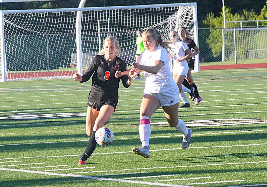 Female soccer players in black and white uniforms compete for the ball near a goal on a sunny field.