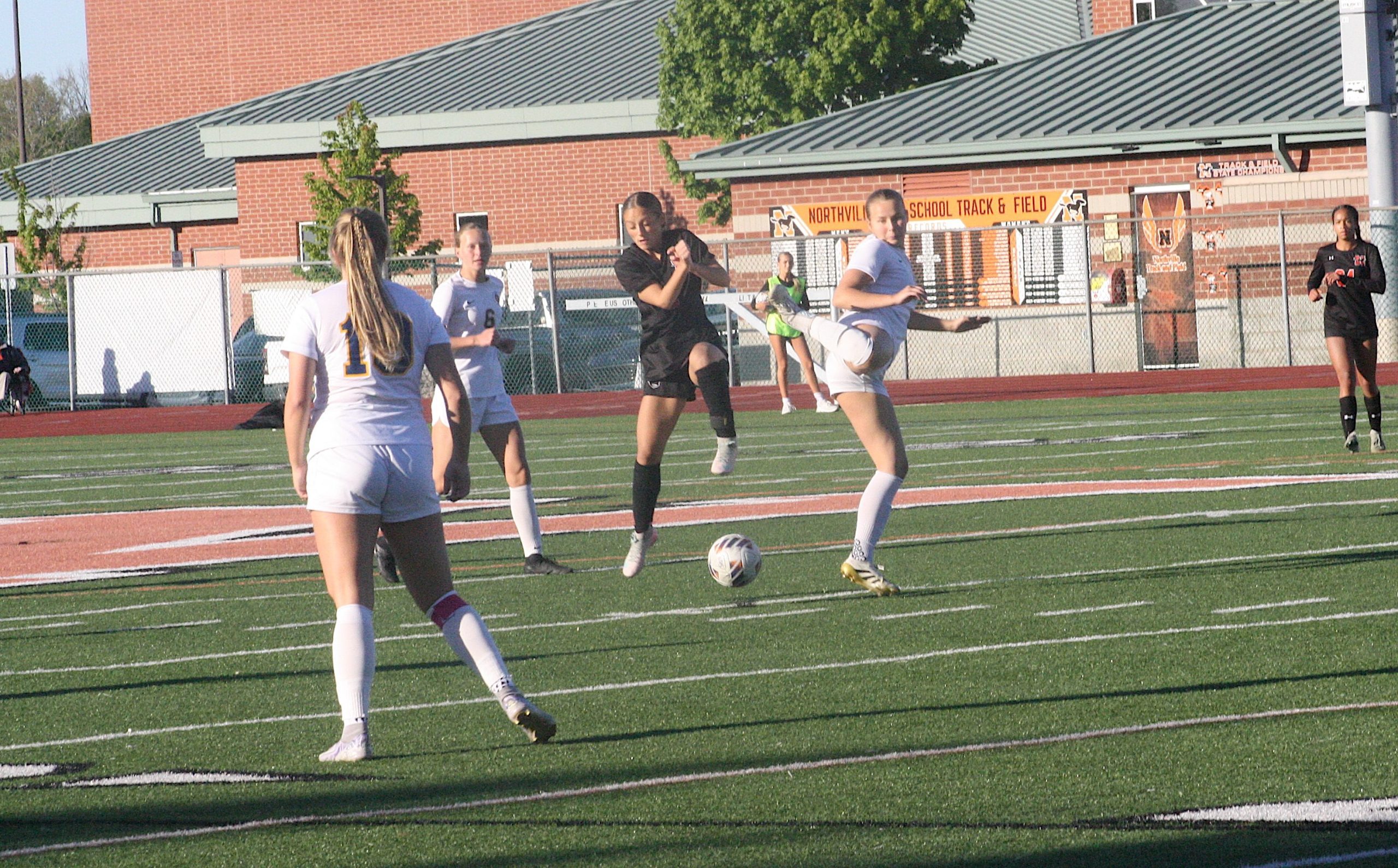 Girls’ soccer game on artificial turf with a ball near the center; brick school building behind.
