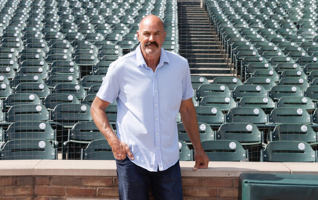 Middle-aged man with a mustache in a light blue short-sleeve shirt stands near empty stadium seats behind a protective net.