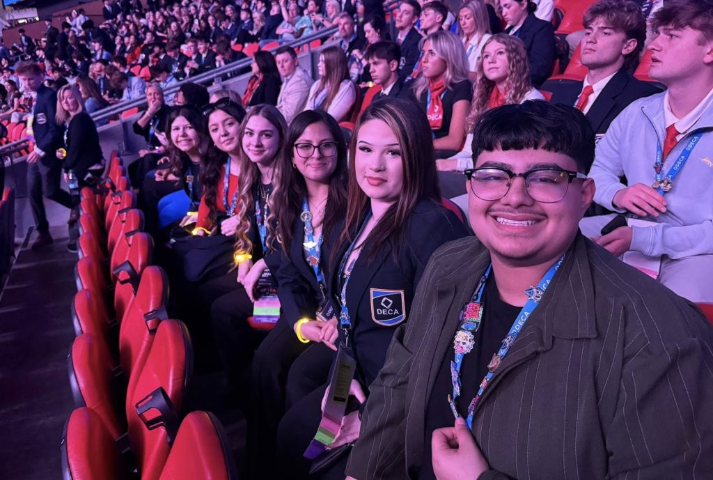 Group of young adults in a stadium seating area taking a selfie, wearing lanyards and badges for a conference/event.
