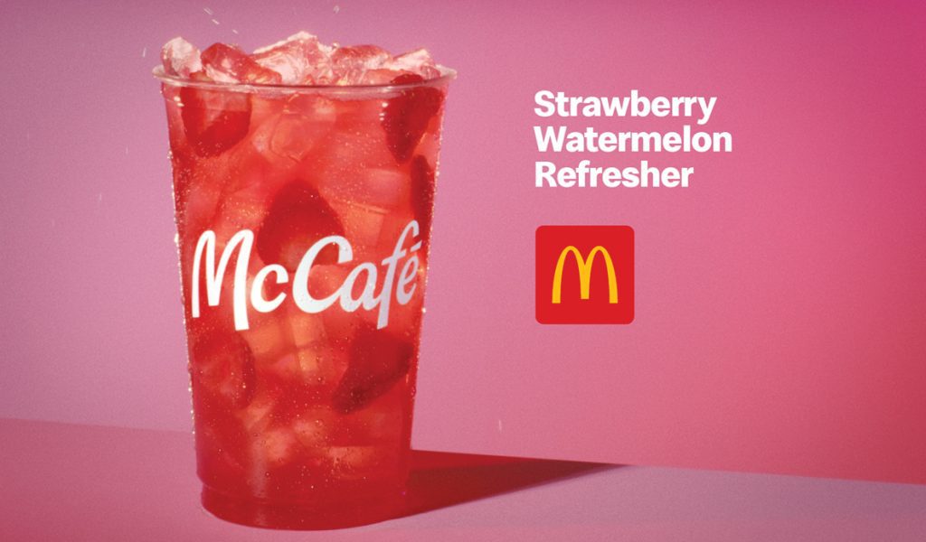 McCafé Strawberry Watermelon Refresher in a red cup with ice, against a pink background with McDonald’s logo.
